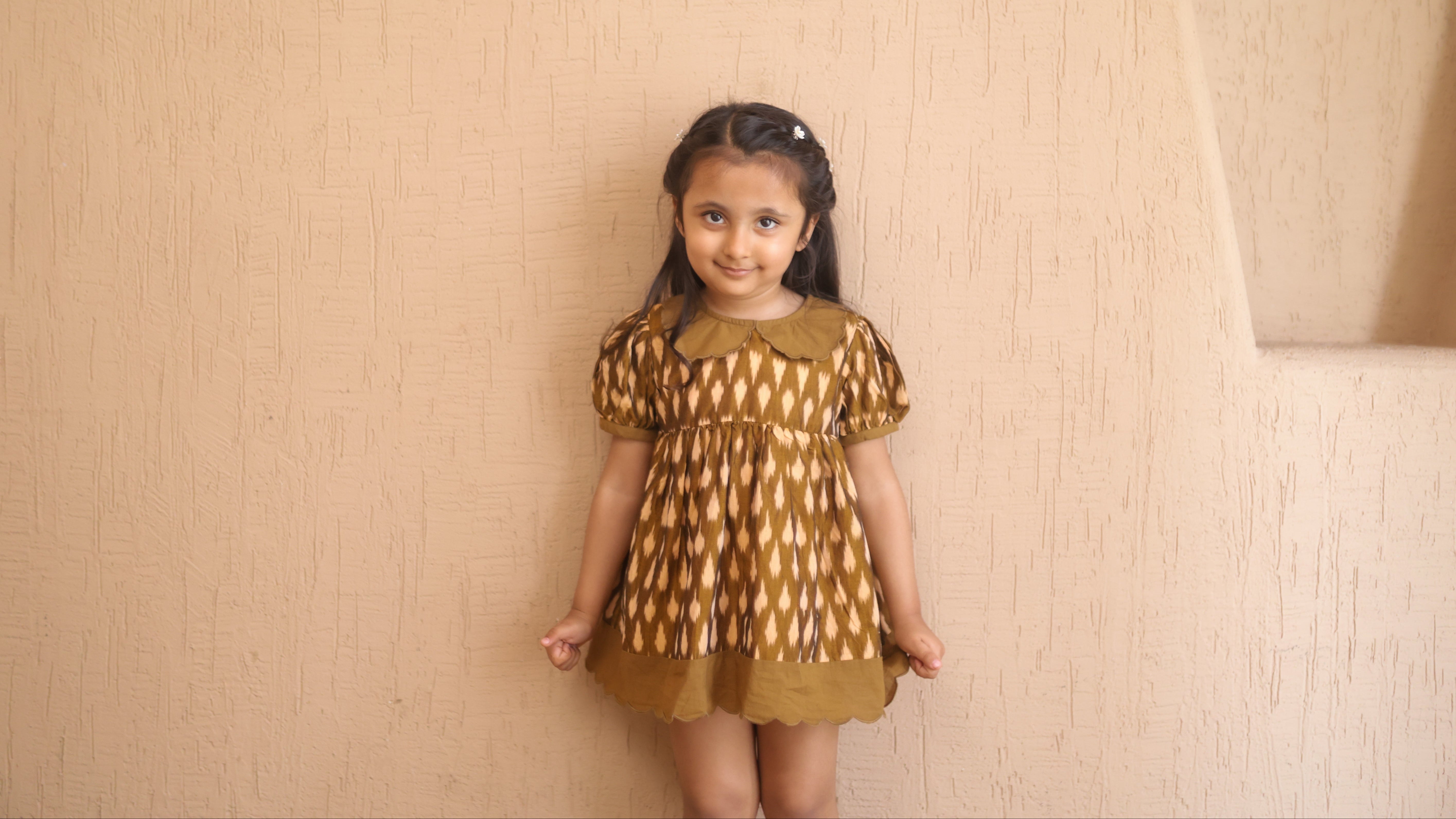 Young girl wearing a patterned dress standing against a beige wall.