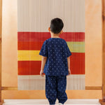 Boy in organic cotton navy block print kidswear stands before colorful woven textile
