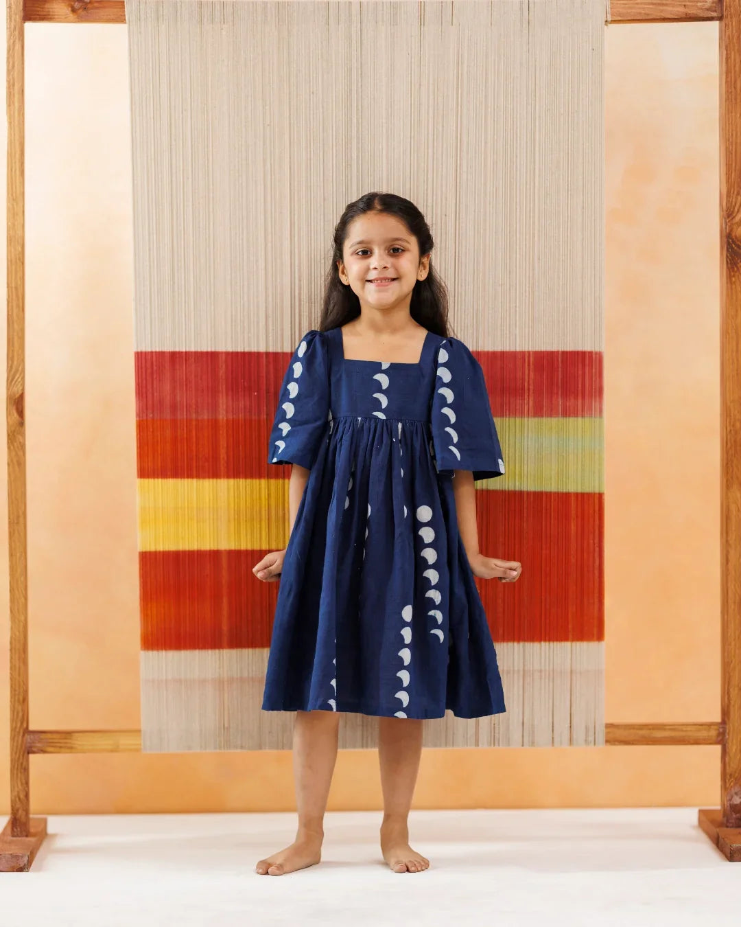 Young girl in a navy blue organic cotton dress with white moon phases print, standing barefoot indoors.
