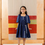 Young girl in a navy blue organic cotton dress with white moon phases print, standing barefoot indoors.