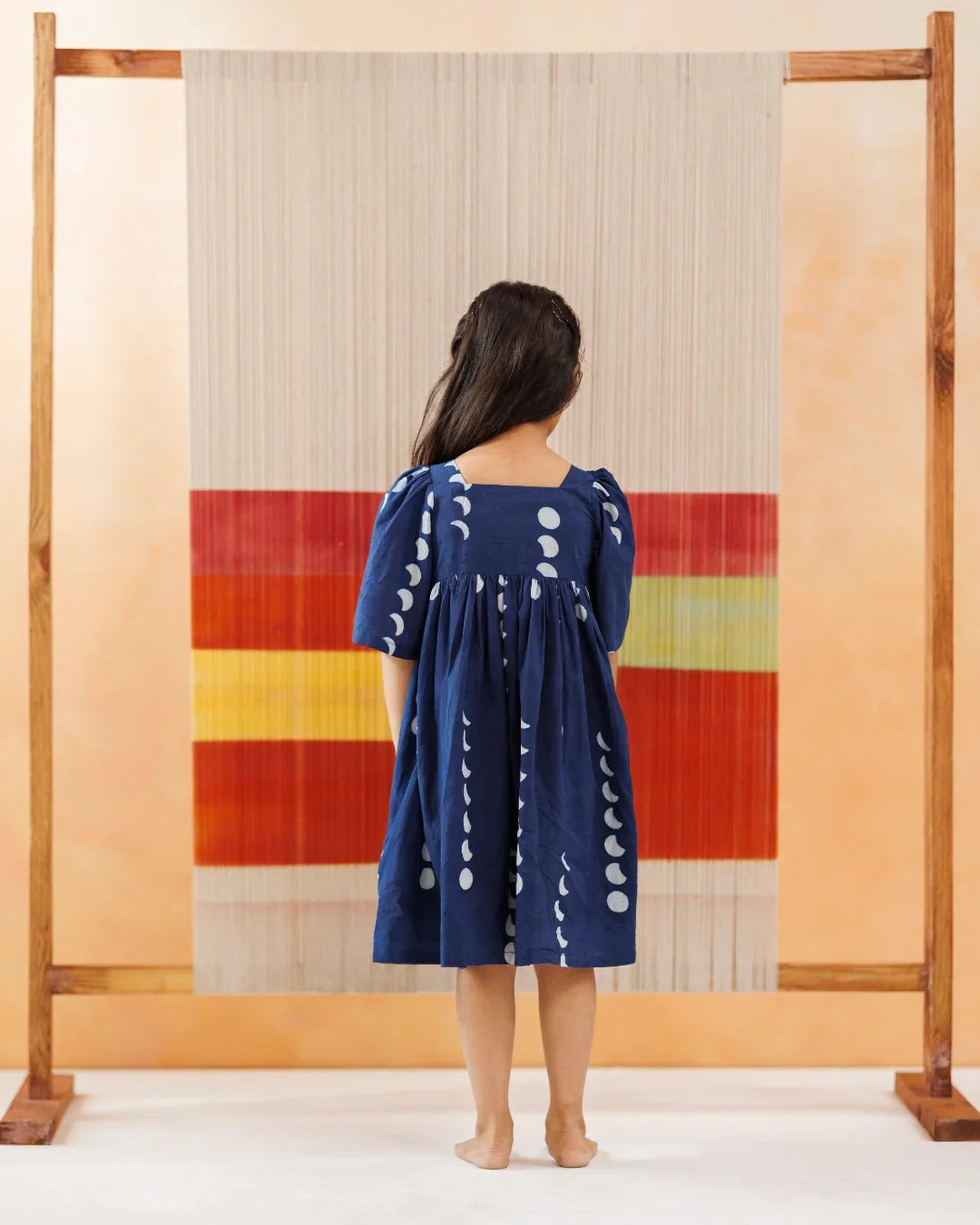 Child in blue organic cotton dress with white block print, standing before a woven textile backdrop