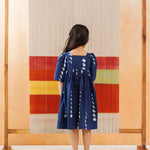 Child in blue organic cotton dress with white block print, standing before a woven textile backdrop