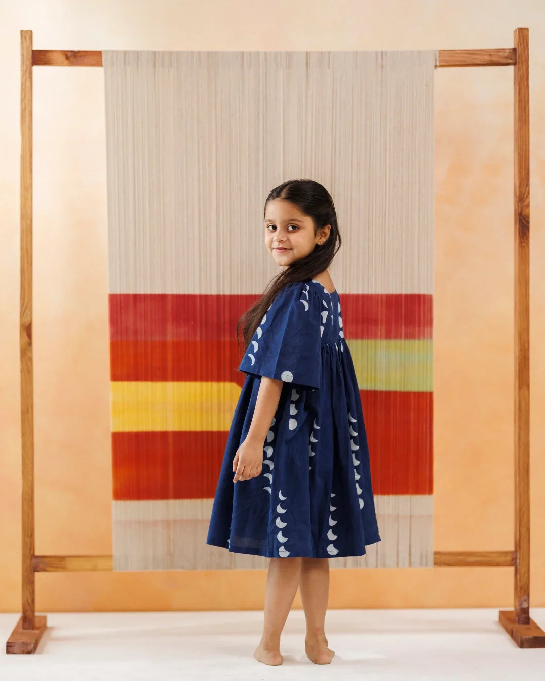 Girl in blue organic cotton block print dress stands barefoot before a colorful textile backdrop