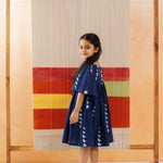 Girl in blue organic cotton block print dress stands barefoot before a colorful textile backdrop