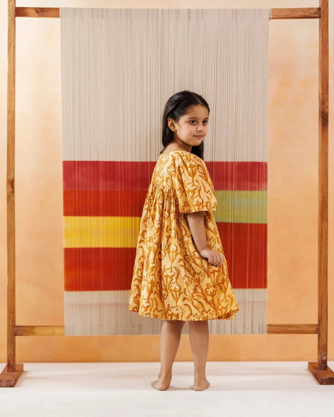 Young girl in yellow floral organic cotton dress standing barefoot by a colorful textile backdrop.