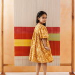 Young girl in yellow floral organic cotton dress standing barefoot by a colorful textile backdrop.