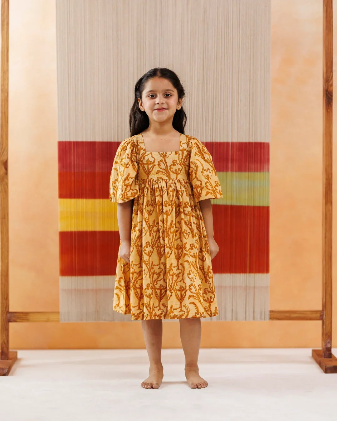 Young girl in a yellow organic cotton block print dress with floral patterns standing indoors