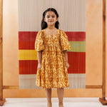 Young girl in a yellow organic cotton block print dress with floral patterns standing indoors
