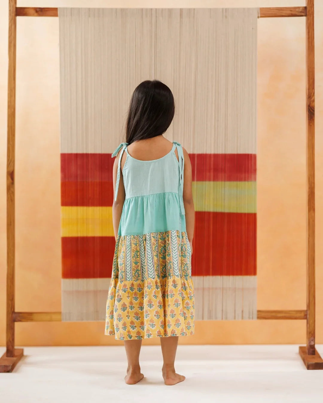 Young girl in a sleeveless pastel tiered dress with block print, standing barefoot indoors