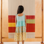 Young girl in a sleeveless pastel tiered dress with block print, standing barefoot indoors