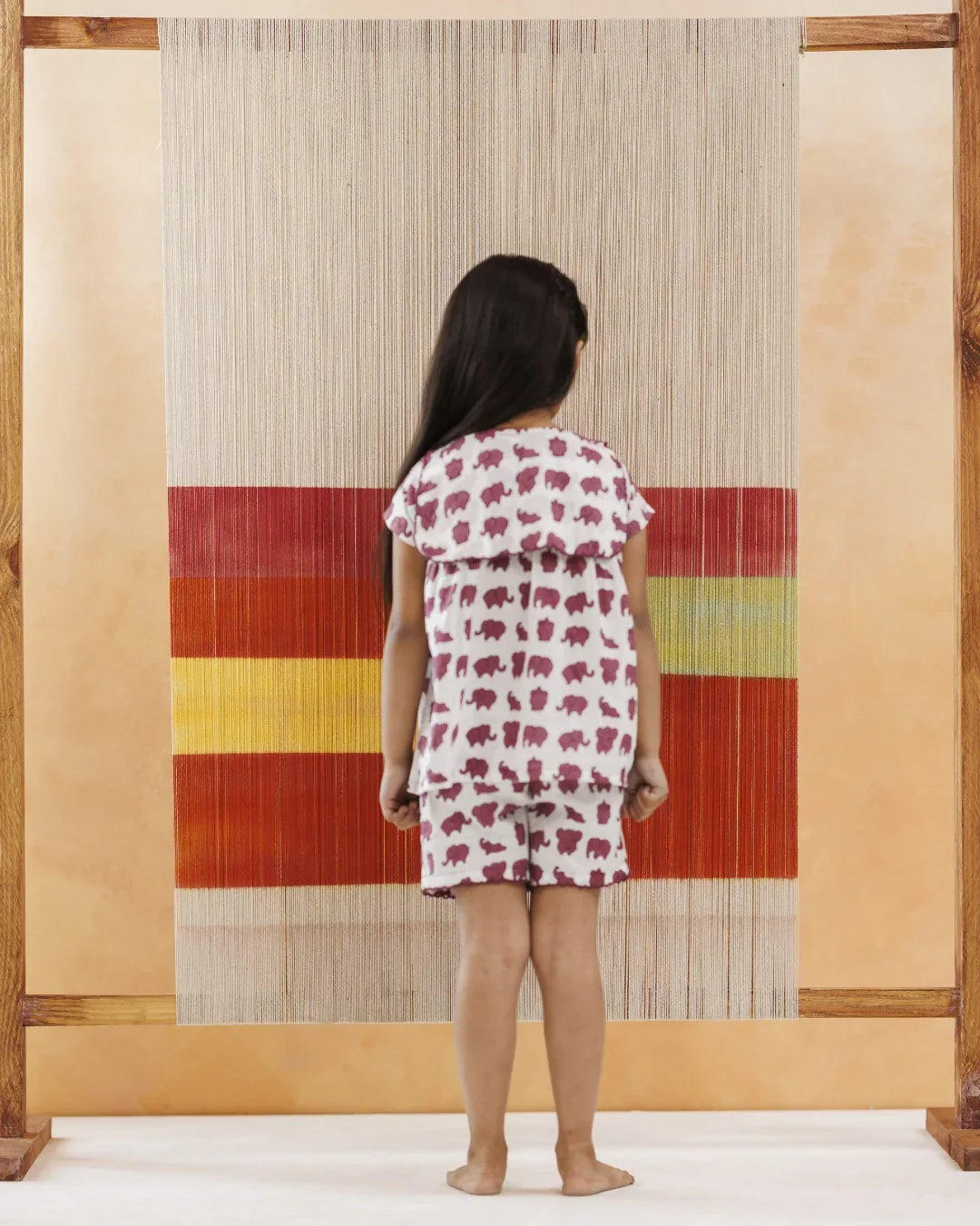 Young child in organic cotton elephant print shorts set, standing indoors by a woven textile backdrop.