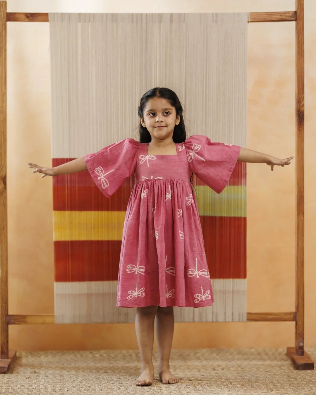 Young girl in a pink organic cotton dress with white dragonfly prints, standing arms outstretched