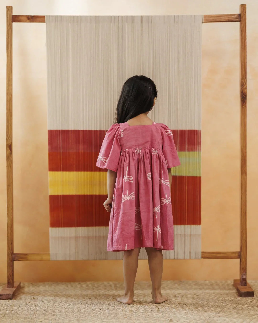 Girl in pink organic cotton dress with dragonfly print standing in front of a woven backdrop