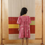Girl in pink organic cotton dress with dragonfly print standing in front of a woven backdrop