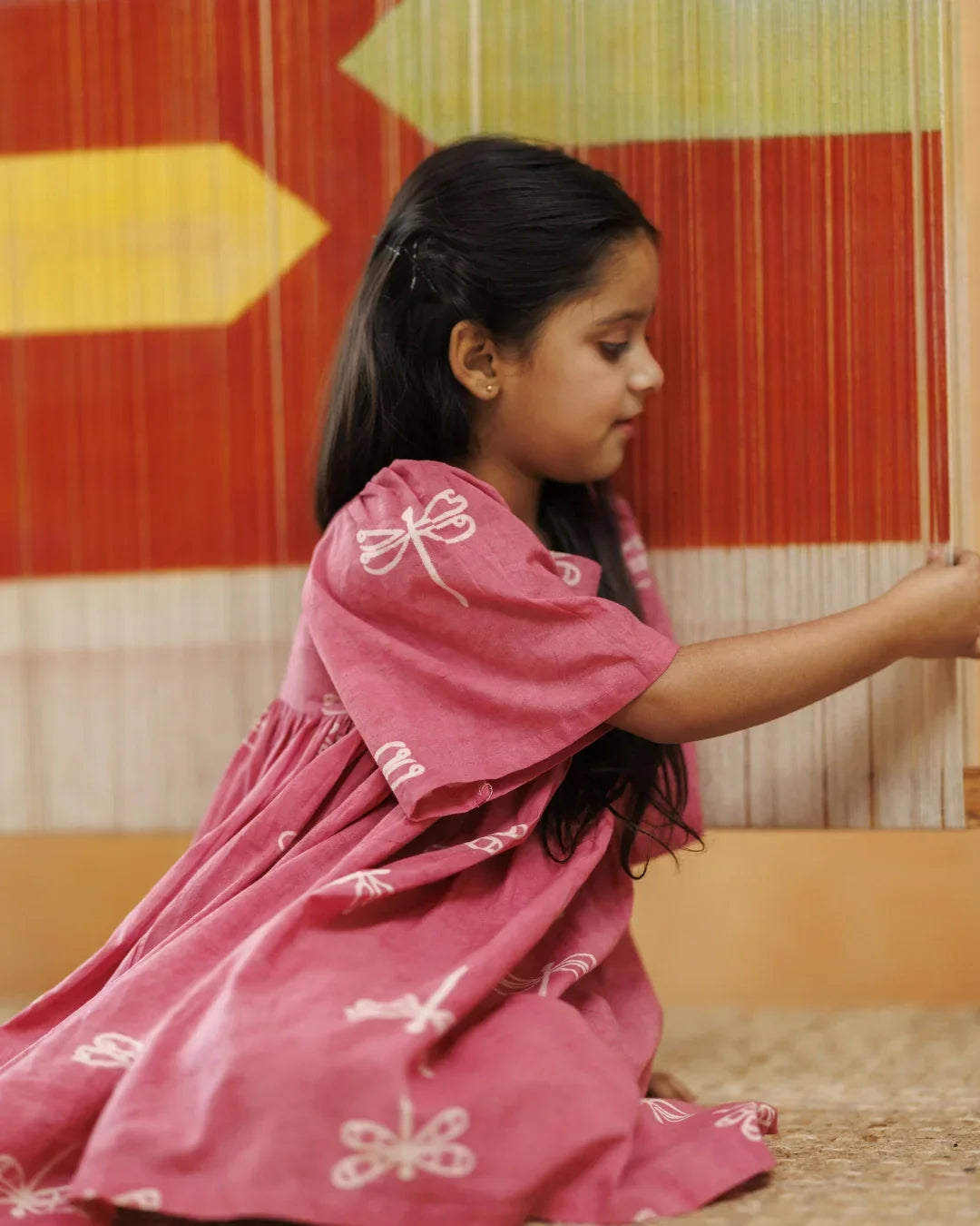 Young girl in organic cotton pink dress with dragonfly print, sitting indoors beside a colorful textile loom.