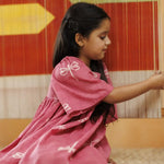 Young girl in organic cotton pink dress with dragonfly print, sitting indoors beside a colorful textile loom.