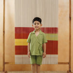 Young boy in green organic cotton kimono top and shorts, standing indoors on woven rug