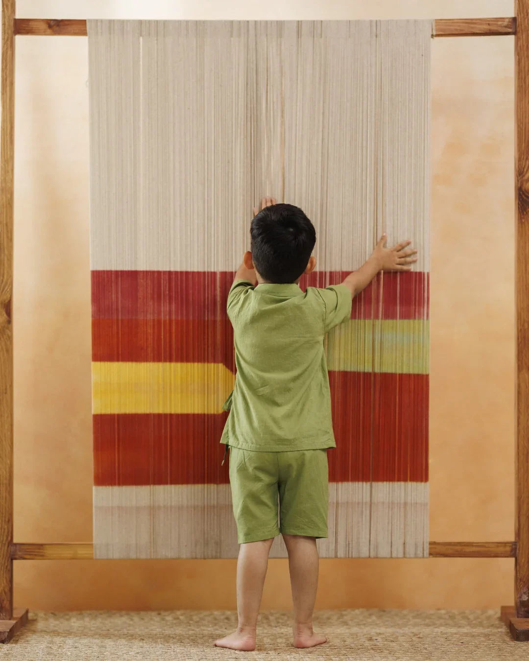 Young child in green organic cotton outfit touching colorful textile loom, sustainable kidswear
