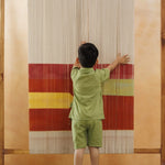 Young child in green organic cotton outfit touching colorful textile loom, sustainable kidswear