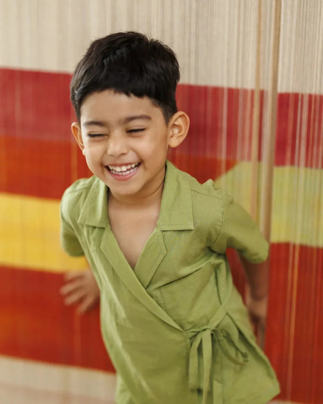 Smiling child in a light green organic cotton wrap top, standing in front of colorful stripes