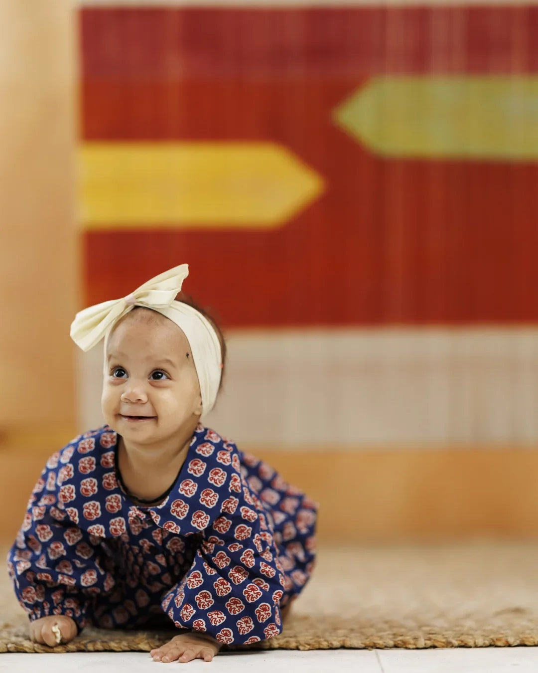 Smiling baby in blue organic cotton block print dress and cream headband crawling on a mat