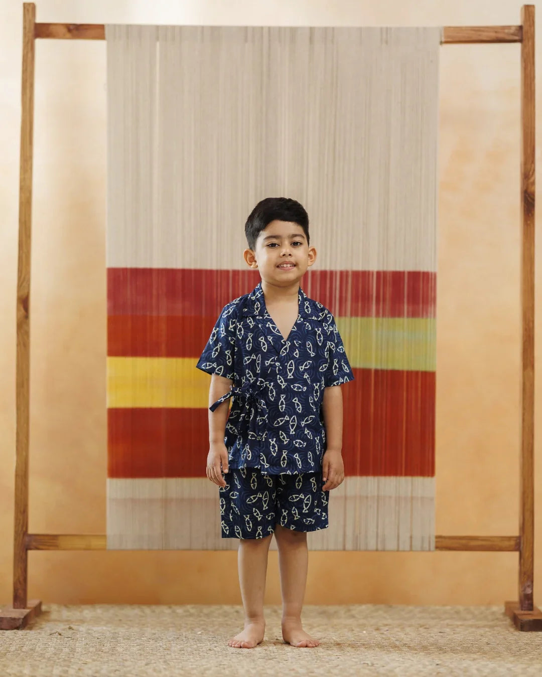 Young boy in blue organic cotton block print shorts set, standing indoors against an ikat textile backdrop
