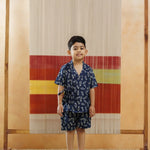 Young boy in blue organic cotton block print shorts set, standing indoors against an ikat textile backdrop