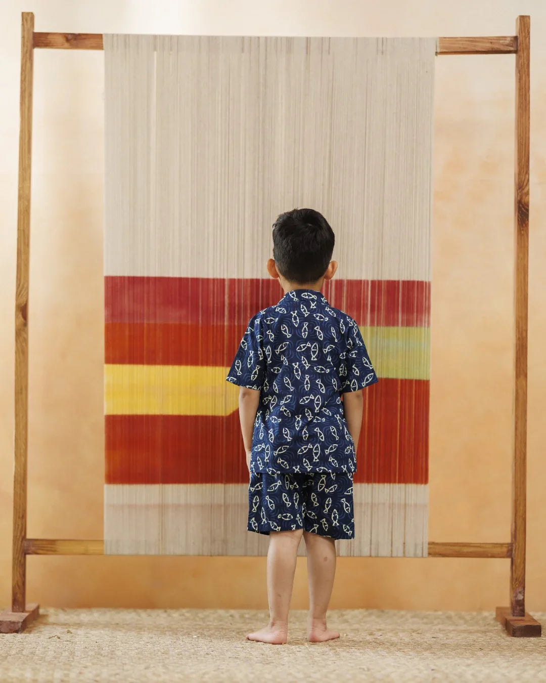 Child in blue organic cotton printed shirt and shorts standing barefoot indoors facing a colorful textile loom
