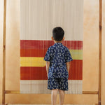 Child in blue organic cotton printed shirt and shorts standing barefoot indoors facing a colorful textile loom
