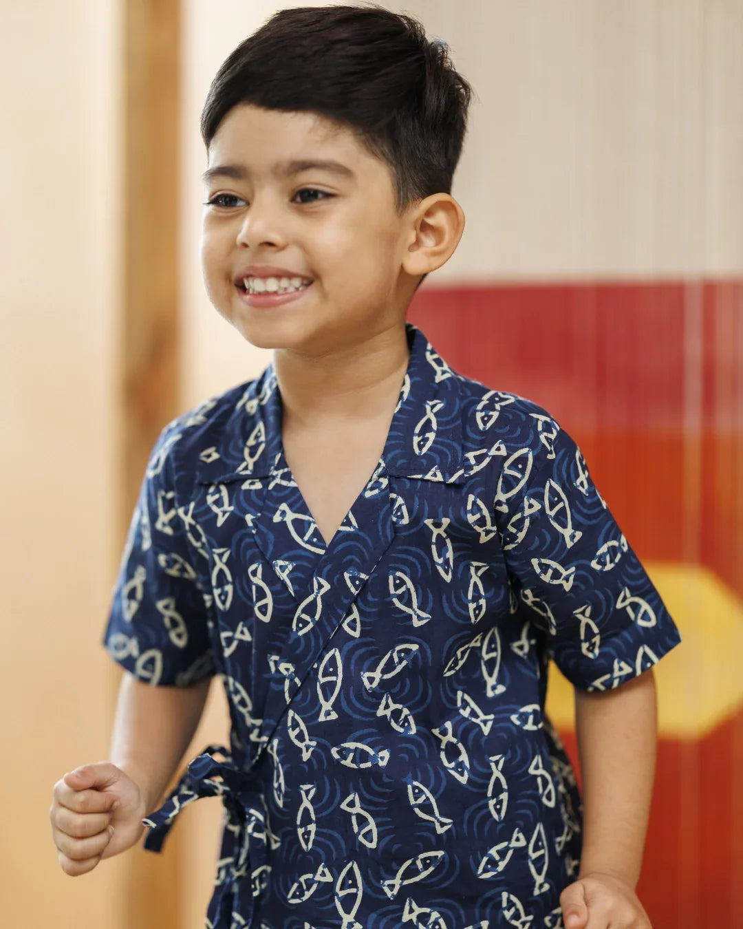 Smiling young boy in blue organic cotton wrap shirt with white fish print, indoor setting