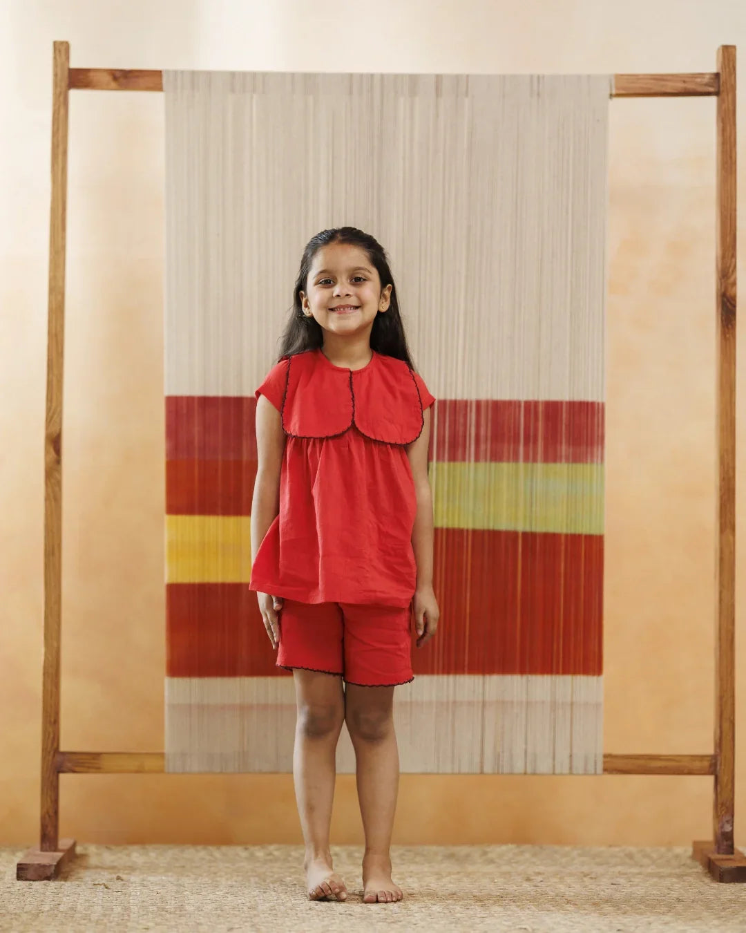 Smiling girl in red organic cotton top and shorts, standing indoors with textile art backdrop.