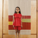 Smiling girl in red organic cotton top and shorts, standing indoors with textile art backdrop.