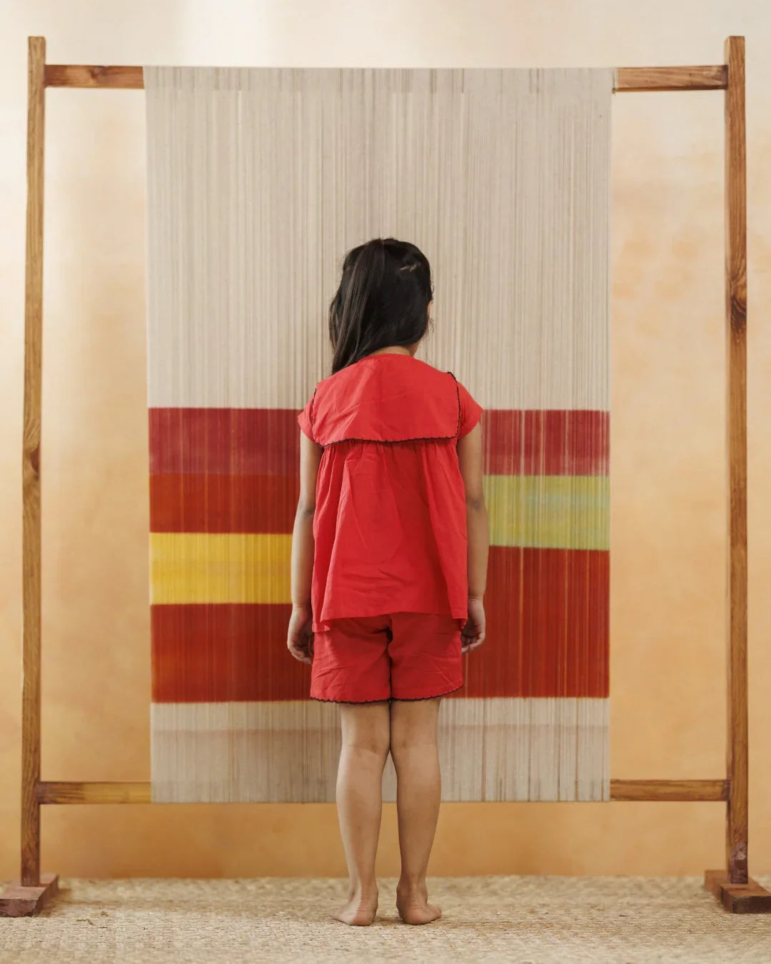 Young child in red organic cotton shorts and top standing barefoot before a colorful woven backdrop.