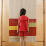 Young child in red organic cotton shorts and top standing barefoot before a colorful woven backdrop.