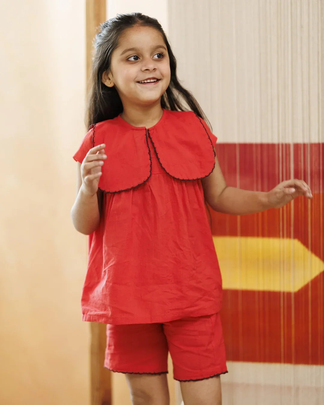 Smiling young girl wearing red organic cotton top and shorts set with scalloped collar indoors