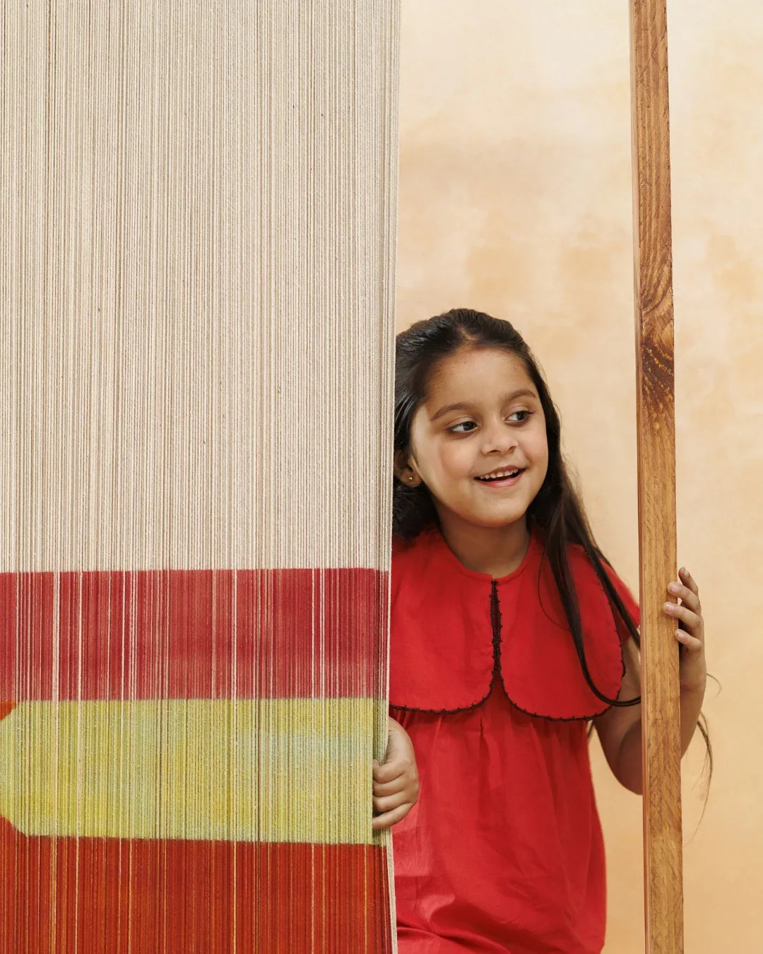 Smiling young girl in a red cotton dress peeking from behind a colorful textile panel