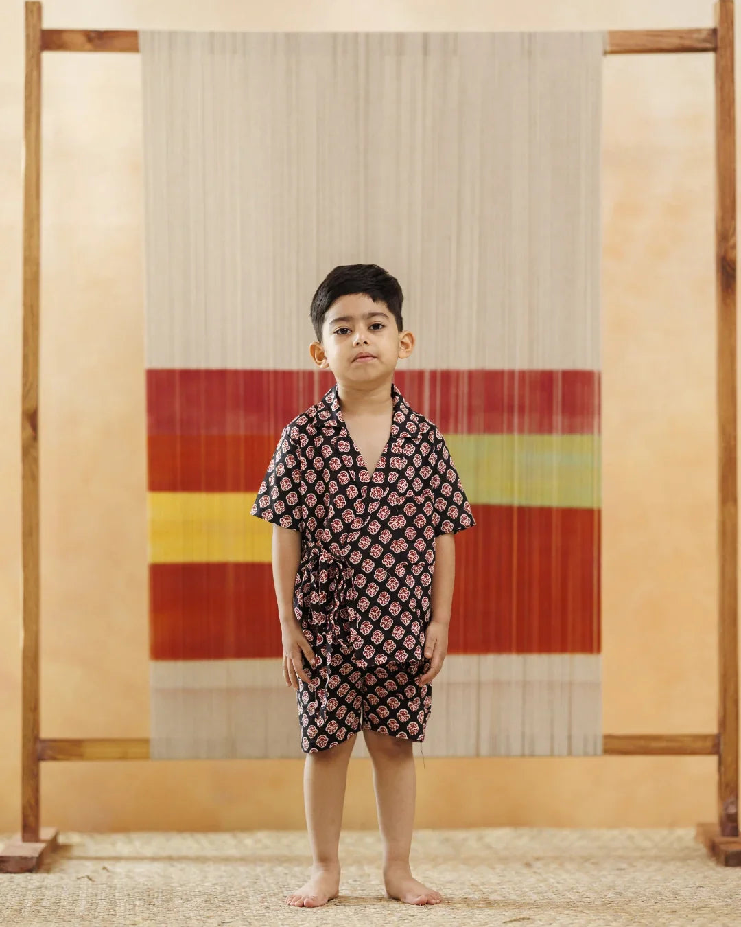 Young boy in organic cotton printed black kimono set stands indoors against a woven textile backdrop.