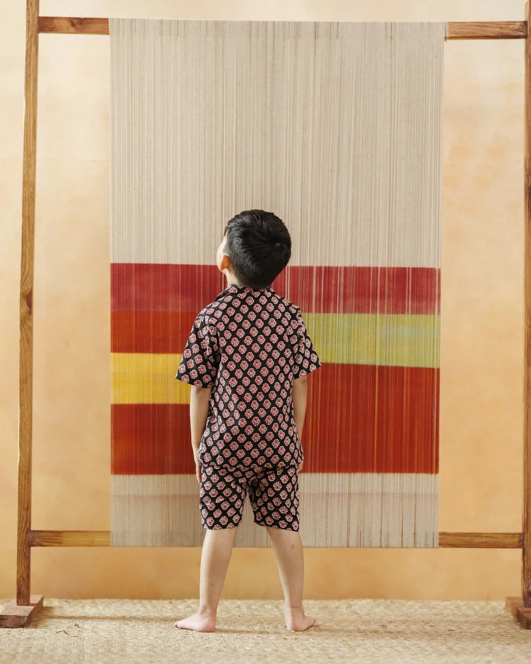 Young boy in organic cotton block print shirt and shorts, facing a colorful woven textile