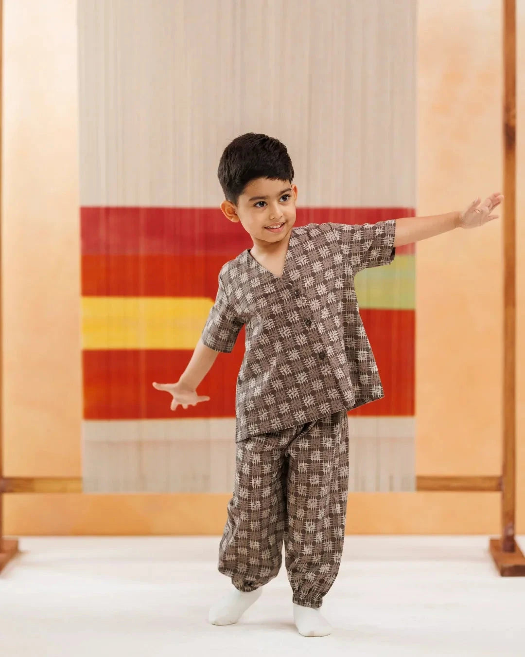 Smiling boy in organic cotton Ikat kidswear set with checked pattern, posing playfully indoors.