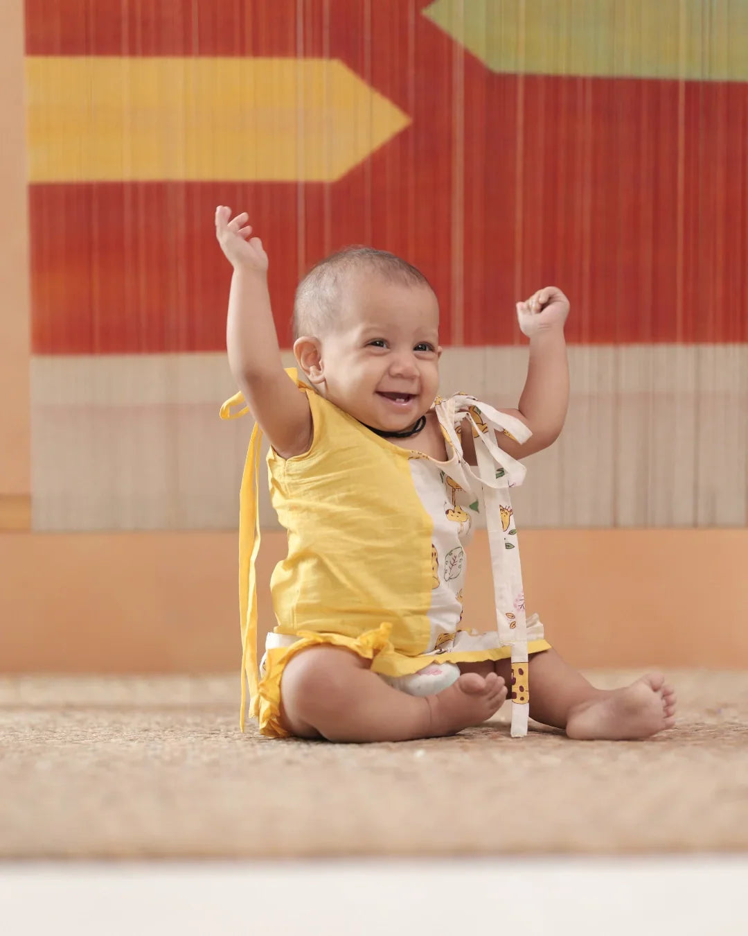 Smiling baby in yellow organic cotton romper sitting on a rug, arms raised, with colorful backdrop