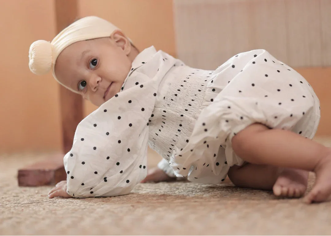 Baby in white organic cotton polka dot romper and headband crawling on carpet