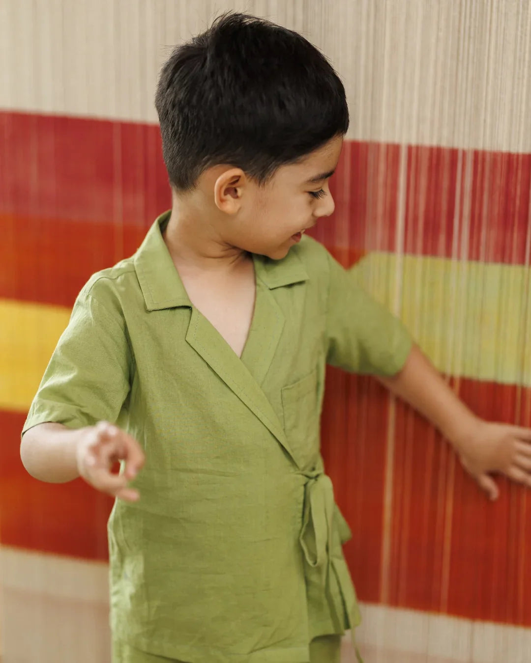Smiling young boy in green organic cotton wrap shirt standing near colorful woven backdrop
