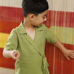 Smiling young boy in green organic cotton wrap shirt standing near colorful woven backdrop