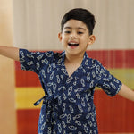 Smiling boy in navy blue organic cotton block print wrap top and shorts, arms outstretched indoors