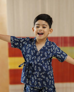 Smiling boy in navy blue organic cotton block print wrap top and shorts, arms outstretched indoors