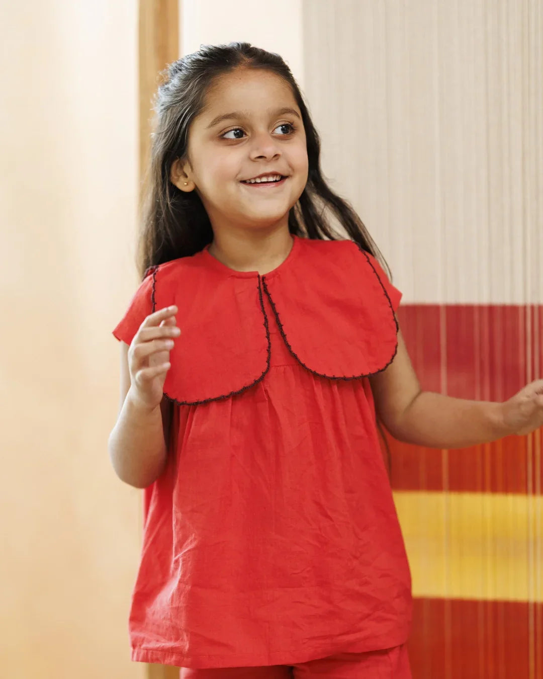 Smiling young girl in a bright red organic cotton top with scalloped collar, indoors